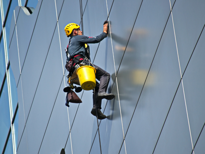 Aggiornamento per addetti ai lavori con sistemi di accesso e posizionamento mediante funi su strutture artificali modulo 1 - AIFOR.IT Worker in a yellow helmet rappelling down the glass exterior of a modern building, with a yellow bucket attached to the harness.