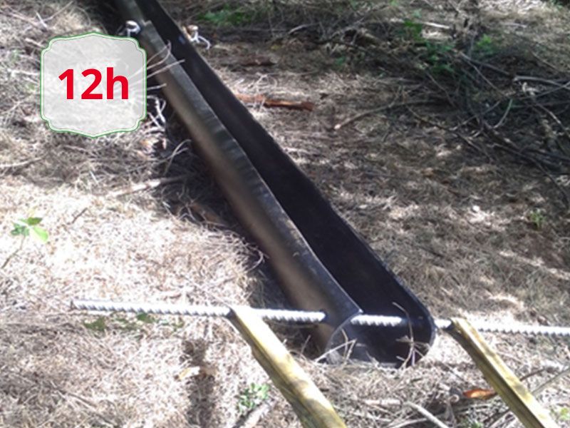 Black metal drainage trough in a dirt trench with wooden stakes nearby; an overlaid '12h' badge is visible.